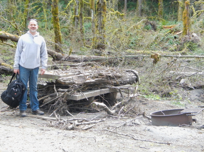 A man stands next to a picnic table covered in woody debris in the Elwha Campground