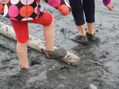 The feet of two young girls are immersed in dark grey Elwha River sediment that looks like clay