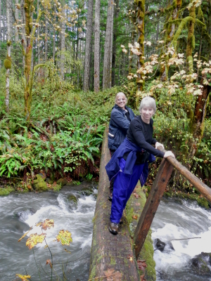 Two smiling hikers crossing a large Elwha River tributary on a narrow bridge with a railing on only one-side of the bridge