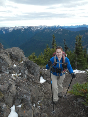 An Olympic Peninsula hiker is smiling as she hikes on a narrow trail with pockets of snow and the Olympic Mountains spread out behind her