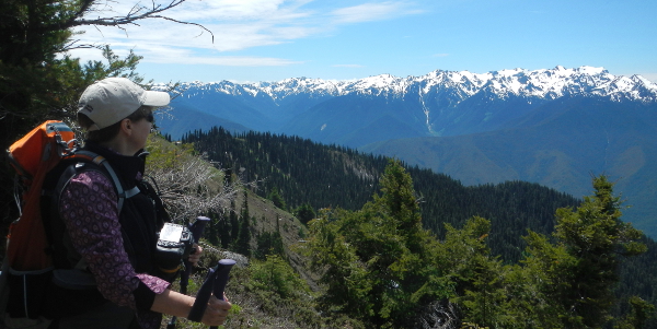 An Olympic Peninsula hiker has her large camera connected to her chest and takes a moment to pose for a photo while enjoying the majestic Olympic Mountains