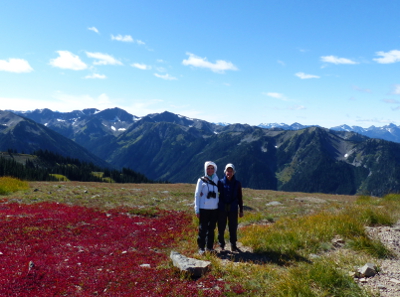 Two birders smiling on Obstruction Point Road at Hurricane Ridge with snow-capped mountains towering in the background