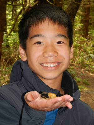 A smiling boy holds up a yellow and black spotted banana slug in the palm of his hand