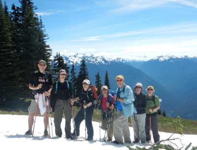 Seven hikers who are decked out in hiking gear pose on the snow at Hurricane Ridge with the mountains in the distance