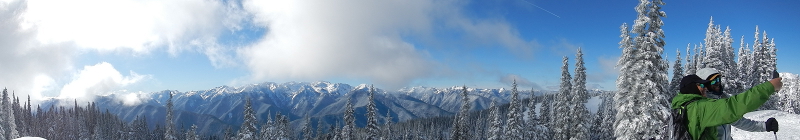 a panorama shows a couple taking a selfie with the Hurricane Ridge snowscape that includes snow covered firs and mountains