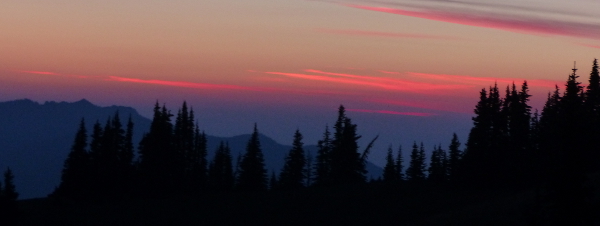 A dark line of trees against a backdrop of blue mountains with red streaks across the sky