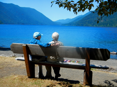 A couple sits on a bench enjoying the stunning view of Lake Crescent and surrounding coniferous forest scenery on a beautiful summer day