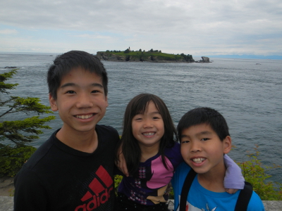 Three siblings stand shoulder to shoulder smiling at the end of the Cape Flattery trail with Tatoosh Island in the background