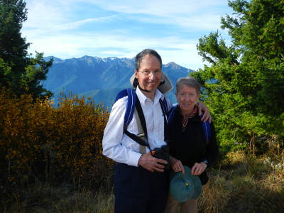 A couple stands smiling on Hurricane Hill with the Olympic Mountains and a brightly colored yellow shrub in the background