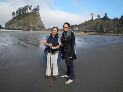 Two hikers stand on the beach smiling at low tide with an expansive sandy beach, small rocky island, and headlands in the background