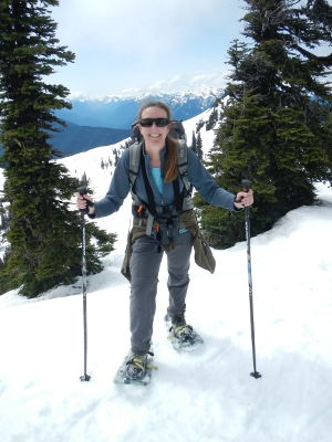Your Olympic Guide is holding ski poles and smiling as she stands on many feet of snow in her snowshoes with the snowclad Olympic mountains in the background