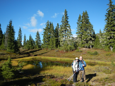 Two hikers stand with a beautiful subalpine lake, subalpine fir, and blue sky behind them