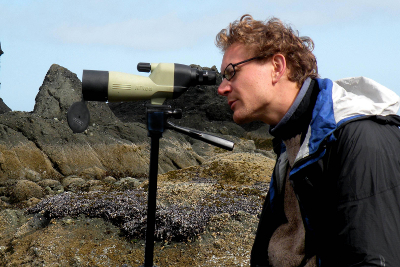 Hiker looking through a spotting scope with rocks and a bed of mussels in the background