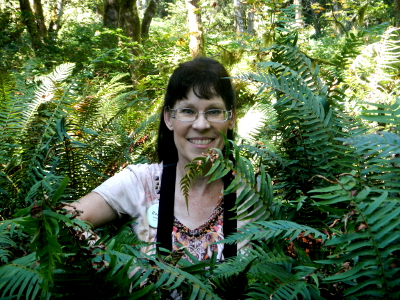 Renee smiling and peering through sword ferns as tall as she is
