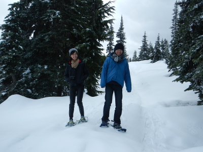 Two smiling hikers tentatively walk on snowshoes through a stand of subalpine fir in the Olympic mountains