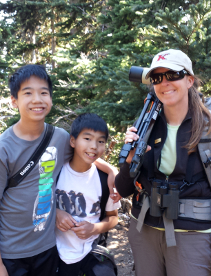 Olympic Guide Carolyn Wilcox stands smiling with her backpack and spotting scope over one shoulder with two young tour participants